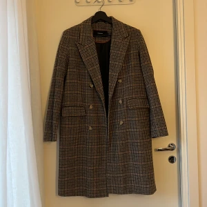 Rutig kappa från Vero Moda, storlek M - Snygg rutig kappa från Vero Moda i klassisk brun, svart och beige färgskala. Dubbelknäppt med stora knappar, bred krage och fickor framtill. Perfekt oversized look med lång ärm och mjukt ullblandat material. En tidlös jacka som lyfter varje outfit.