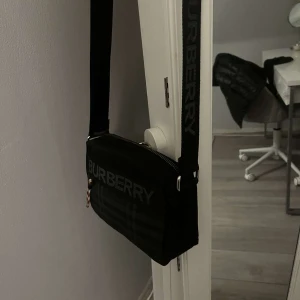 Svart axelväska från Burberry - Snygg svart axelväska från Burberry med klassiskt rutmönster i mörka toner och stort logotypband framtill. Väskan har justerbar axelrem med Burberry-tryck och dragkedja med färgglad detalj. Perfekt storlek för det viktigaste och riktigt stilren design.