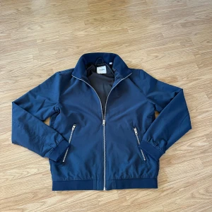 Blå vindjacka från Jack & Jones S - Snygg marinblå vindjacka från Jack & Jones med dragkedja framtill och två fickor med dragkedja. Jackan har hög krage och ribbade muddar vid ärmslut och midja. Perfekt för dig som gillar stilren och enkel design.