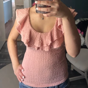 Rosa volangblus med struktur - Supersöt ljusrosa blus med volangdetaljer runt ringningen och axlarna. Toppen är gjord i ett krinklat, strukturerat tyg. 