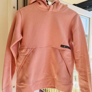 Rosa hoodie från Sail Racing - Säljer en snygg rosa hoodie från Sail Racing med huva och snörning. Tröjan har en stor magficka och en diskret logga på framsidan. Mjuk och skön i bomullsmix, perfekt för chill dagar. Passar dig som gillar stilren och enkel design. Knappt använd och i mycket gott skick.
