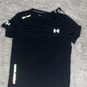 Svart Under Armour Threadborne t-shirt - Svart t-shirt från Under Armour Threadborne med reflekterande logga och textdetaljer på bröstet, ärmen och nedtill. Tillverkad i ett lätt och tekniskt polyestermaterial som andas, perfekt för träning. Klassisk passform med korta ärmar och rund hals.
