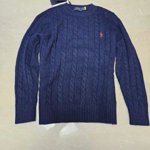 Marinblå tröja Polo Ralph Lauren - Säljer en snygg mörkblå stickad tröja från Ralph Lauren med det klassiska röda logotypbroderiet på bröstet. Tröjan har ett kabelstickat mönster och långa ärmar med ribbade muddar. Perfekt för kyligare dagar. Priset kan diskuteras vid snabb affär.