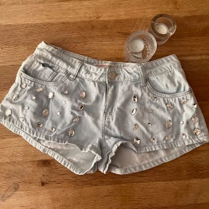 Ljusblå jeansshorts med diamanter  - Snygga ljusblå jeansshorts från Toxik3 med coola paljett- och sten-detaljer framtill. Klassisk femficksmodell med bälteshällor och knappstängning. Perfekta för sommarens alla äventyr och ger en riktigt trendig vibe. Finns ingen storlek skriven men skulle gissa runt xs till m