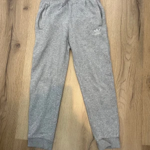 Grå mjukisbyxor från Adidas - Gråa mjukisbyxor från Adidas med klassisk logga broderad på ena benet. Byxorna har resår i midjan med snörning, två sidofickor och muddar vid bensluten. Perfekta för chill eller träning, riktigt sköna och stilrena.