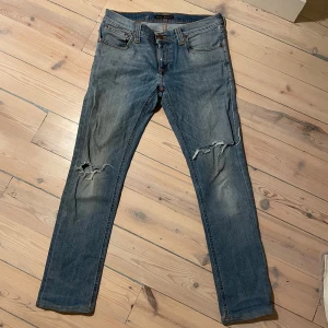Nudie Jeans blå slitna jeans W30 L34 - Säljer blå Nudie jeans som är förkortade, jeansen är ungefär 90 cm långa från midjan till foten. Jeansen har slitningar och hål på båda knäna. Klassisk femficksmodell med orangea sömmar och läderpatch bak. Jeansen har en rak passform och är tillverkade i bomull.