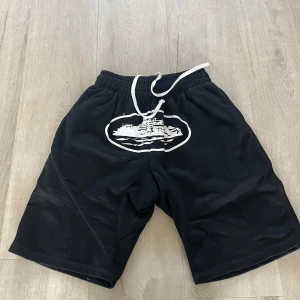 Svarta shorts från corteiz - Svarta corteiz shorts med vitt tryck framtill och vit snörning i midjan. Modellen är loose fit med elastisk midja och bakficka med knapp. Perfekta för chill dagar eller träning. Materialet känns som mjuk bomull eller syntet.