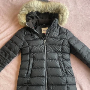 Svart pufferjacka från Tommy Jeans - Säljer en svart pufferjacka från Tommy Hilfiger med fluffig fuskpäls på huvan. Har inga stora tecken från användingen.