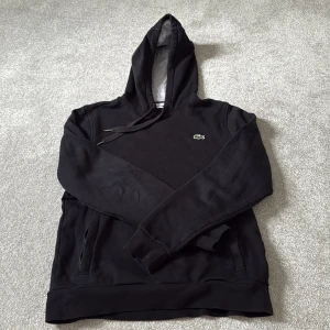 Svart hoodie från Lacoste Sport - Svart hoodie från Lacoste Sport med klassisk krokodillogga på bröstet. Tröjan har huva med snörning, långa ärmar och fickor på sidorna. Insidan av huvan är grå. Perfekt för chill dagar eller när du vill ha en sportig look.