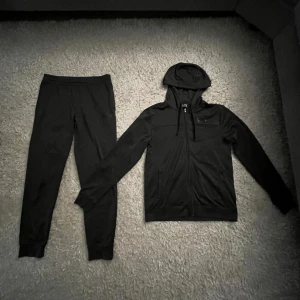 Svart hoodie från EA7 Emporio Armani - Svart hoodie från EA7 Emporio Armani med dragkedja och huva. Tröjan har långa ärmar, snörning vid huvan och diskret logga på bröstet. Tillverkad i mjukt och lätt material, perfekt för chill eller träning.