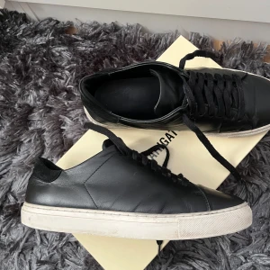 Svarta sneakers från Axel Arigato - Stilrena svarta sneakers från Axel Arigato i skinn med klassisk vit platt sula.  passar till det mesta. Perfekta för dig som gillar en clean och modern look. Skorna är stora i storleken finns en liten skada i insida hälen men inget som märks vid användning se på sista bilden! Finns täcken på användning men fortfarande fina