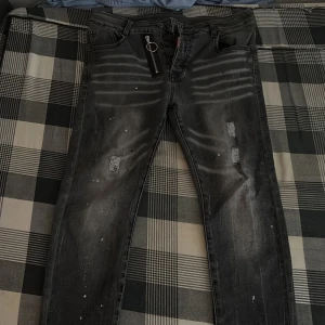 Svarta Dsquared2 jeans med dragkedja - Säljer ett par svarta jeans från Dsquared2 med slitningar, färgstänk och en snygg silverfärgad dragkedja med ringdetalj framtill. Jeansen har smal passform och klassisk femficksdesign. Perfekta för dig som gillar streetstyle och edgy detaljer.