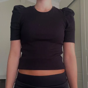 Svart ribbad topp med puffärm - Svart ribbad topp med korta puffärmar och rund halsringning. Modellen är croppad och sitter tight mot kroppen, vilket ger en snygg siluett. Använd 1 gång!