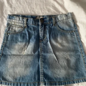 Blå jeanskjol med slitningar - Snygg blå jeanskjol i kort modell med slitna detaljer framtill och coola bakfickor med knappar och liten röd etikett. Klassisk femficksdesign och bälteshällor. Perfekt för dig som gillar en avslappnad och trendig stil. Storlek 146 men passar även xs-s 