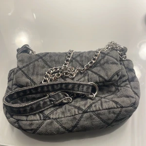 Quiltad grå axelväska med kedja - Säljer en snygg quiltad axelväska i grått jeansmaterial med silverfärgad kedjerem. Väskan har ett rutigt mönster och en mjuk, puffig form som ger en cool vibe. Perfekt storlek för det viktigaste och passar till många outfits.