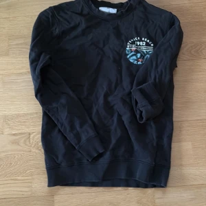 Svart sweatshirt från DNMD Venice Beach - Svart långärmad sweatshirt från DNMD med tryck på bröstet där det står 'Venice Beach 1982'. Tröjan har rund halsringning och ribbade muddar vid ärmslut och nederkant. Perfekt för en chill och avslappnad stil.