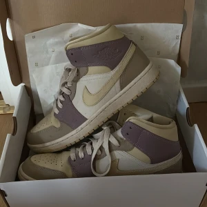 Nike Air Jordan 1 i beige och lila - Nike Air Jordan 1 sneakers i en snygg mix av beige, ljusgul och lila toner. Skorna har klassisk snörning, rund tå och platt sula. Tillverkade i skinn med ikoniska Jordan-detaljer på sidan och hälen. Perfekta för dig som vill sticka ut med stilrena färgblock.