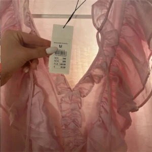 Rosa volangklänning från Kappahl - Säljer en söt ljusrosa kortklänning från Kappahl med volanger längs v-ringningen och axlarna. Klänningen är ärmlös och har ett skirt, glansigt material som ger en romantisk vibe. Perfekt för dig som gillar feminina detaljer och en lätt känsla.