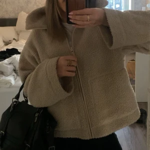 Beige teddyjacka med dragkedja - Supermjuk och fluffig teddyjacka i beige med hög krage och hel dragkedja framtill. Jackan har två stora fickor och en avslappnad passform som är perfekt för kyliga dagar. Passar dig som gillar en cozy och trendig look. Jackan passar mig som vanligtvis har storlek XS/S/M