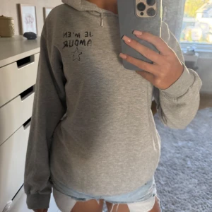 Grå hoodie från Gina Tricot med tryck - Grå hoodie från Gina Tricot med svart texttryck 'Je m'en amour' och en stjärna både fram och bak. Mjuk insida, huva och långa ärmar. 