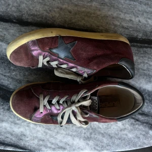 Vinröda sneakers från Golden Goose - Snygga vinröda sneakers från Golden Goose med svart stjärndetalj på sidan och lilarosa metallicinslag. Skorna har grå snörning, rund tå och en klassisk platt sula. Ovandelen är i mocka och detaljer i skinn, perfekt för en cool vardagslook. de är dock i ganska använt skick men det är inget som märks i användningen eller på utseendet på längre håll:) perfekta höstskor