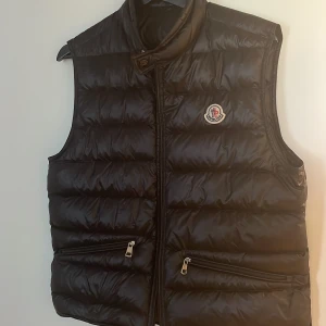 Svart dunväst från Moncler - Snygg svart dunväst från Moncler med glansig finish och klassisk logga på bröstet. Västen har två dragkedjefickor framtill och hög krage. Perfekt att slänga över en hoodie eller tröja för extra värme och stilpoäng.
