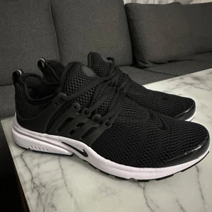 Nike Presto svarta sneakers - Svarta Nike Presto sneakers med vit sula och mesh-ovandel för extra andningsförmåga. Snygg design med svarta detaljer, snörning och diskret Nike-logga på sidan. Perfekta för dig som gillar sportig och clean stil.