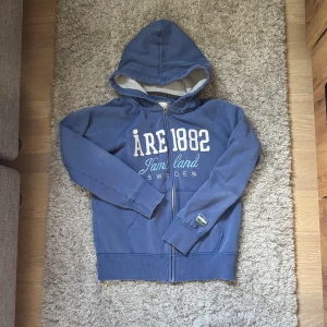 Vintage Åre Hoodie - Blå vintage Åre hoodie med stort vitt tryck 'ÅRE 1882 Jämtland Sweden' på bröstet Den har ett jättelitet hål på ryggen. Tröjan har huva med snörning, dragkedja framtill och två fickor. Perfekt för dig som gillar chill och sportig stil.