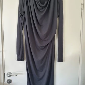 Grå långklänning från Day Birger et Mikkelsen - Snygg grå långklänning med draperad halsringning från Day Birger et Mikkelsen. Klänningen har lång ärm och är tillverkad i ett mjukt, stretchigt material som faller snyggt längs kroppen. Perfekt för dig som gillar minimalistisk och stilren design.