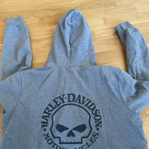 Grå Harley-Davidson hoodie XL - Grå hoodie från Harley-Davidson i storlek XL med dragkedja och huva. Stor svart dödskalle-logga på ryggen och mindre logga på bröstet. Mjuk bomullsblandning och känguruficka framtill. Perfekt för dig som gillar streetwear och motorcykelstil.