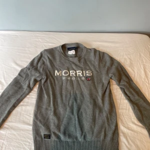 Grå stickad tröja från Morris - Snygg grå stickad tröja från Morris Rowing Club med broderad logga och brittisk flagga framtill. Tröjan har rund hals, långa ärmar och bred ribbad mudd vid nederkant och ärmslut. Perfekt för dig som gillar klassisk och stilren look. Pris kan diskuteras. 