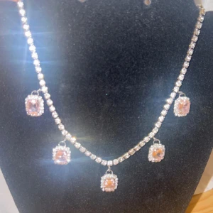 Glittrigt halsband med rosa stenar - Säljer ett glittrigt halsband med fem hängande fyrkantiga rosa stenar, omgivna av små klara stenar. Kedjan är täckt av gnistrande stenar i silverfärg. Perfekt för dig som vill sticka ut med lite extra bling.