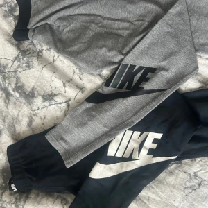 Nike träningsbyxor svart och grå - Två par mjukisbyxor från Nike, ena i grått med svart logga och den andra i svart med vit logga. Båda har elastiska muddar vid benslut och är gjorda i mjukt bomullsmaterial. Perfekta för chill eller träning, med stor Nike-print längs benet. 100kr/st