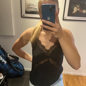 Svart linnetopp med spetsdetaljer - Säljer en svart linnetopp med tunna axelband och snygga spetsdetaljer längs v-ringningen och under bysten. Toppen har korsade band i ryggen. Endast använd 1gång🥰