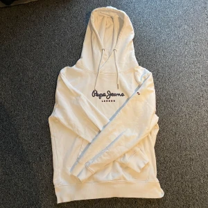 Vit hoodie från Pepe Jeans London - Säljer en stilren vit hoodie från Pepe Jeans London med svart broderad logga på bröstet. Tröjan har huva med snörning, känguruficka och ribbade muddar. Perfekt för dig som gillar en clean och klassisk look. Bra skick, stl m och passar dig runt 180! Bara att höra av sig vid frågor eller funderingar.