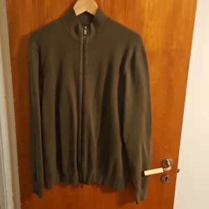 Olivgrön kofta från Hampton Republic - Olivgrön kofta från Hampton Republic i storlek M. Tröjan är stickad i bomull, har hel dragkedja framtill och hög krage. Perfekt för lager-på-lager och enkel att matcha med jeans eller chinos. Långärmad och stilren design.