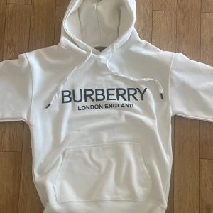 Vit Burberry hoodie med logga - Snygg vit hoodie från Brbry med stor svart logga på bröstet och texten 'London England'. Tröjan har huva med snörning och en klassisk känguruficka framtill. Perfekt för dig som gillar stilrena och exklusiva plagg. HELT OANVÄND 