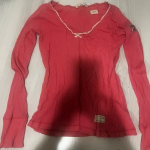 Rosa långärmad topp från Smoglia - Supersöt rosa långärmad topp från odd molly med ribbad struktur och v-ringad hals med spetskant och liten rosett. På ena ärmen finns ett coolt hjärtmönster med stjärna och vingar som defenerar odd Molly märket. 