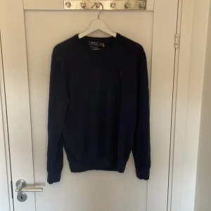 Mörkblå merinoulltröja Polo Ralph Lauren - Snygg mörkblå tröja från Polo Ralph Lauren i mjuk och tvättbar merinoull. Klassisk rund hals och långärmad modell med diskret logga på bröstet. Perfekt för dig som gillar stilrena och tidlösa plagg med premiumkänsla. Storlek L passar även M