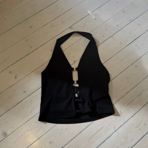 Svart omlottväst med metallspännen - Snygg svart väst med halterneck och djup v-ringning. Framsidan har två coola metallspännen som håller ihop västen och ger en edgy look. Västen är croppad och har en omlottkänsla framtill. Perfekt för att lyfta vilken outfit som helst.