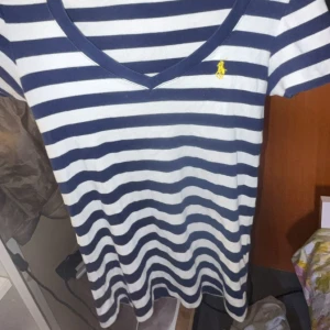 Randig t-shirt från Ralph Lauren - Snygg t-shirt från Ralph Lauren med marinblå och vita ränder, klassisk v-ringning och korta ärmar. Broderad gul logga på bröstet. Skön bomullskvalitet och perfekt passform för en avslappnad stil.
