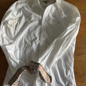 Vit Burberry skjorta med rutig detalj - Vit skjorta från Burberry i slim fit med klassisk krage och knappar framtill. Insidan av kragen och manschetterna har den ikoniska Burberry-rutiga detaljen i beige, svart och rött. Skjortan är långärmad och tillverkad i mjuk bomull.  Lite gul i armhålan men inget som syns jätte mycket