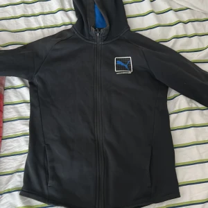 Svart hoodie från Puma med dragkedja.  - Svart hoodie från Puma med hel dragkedja och huva. Tröjan har ett blått Puma-logomärke på bröstet och är fodrad med blått meshtyg på insidan av huvan. Perfekt för dig som gillar sportig och clean stil.