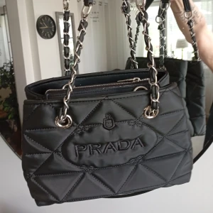 Svart quiltad handväska  - Snygg svart handväska med quiltad design och silverfärgade kedjedetaljer på axelremmen. Väskan har ett klassiskt triangelformat P-emblem framtill och broderad logga på baksidan. Perfekt storlek för det viktigaste och har dragkedja upptill.