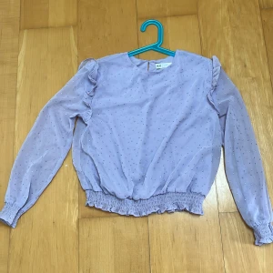 Ljuslila blus med volang från H&M - Supersöt ljuslila blus från H&M med små glittriga prickar över hela tyget. Blusen har långa ärmar, volangdetaljer på axlarna och smockad nederkant samt ärmslut. Perfekt för dig som vill ha något extra fint och trendigt!