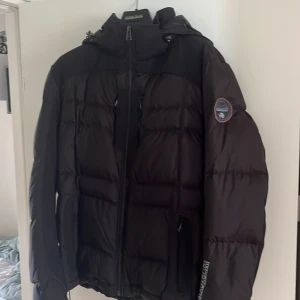 Napapijri SKIDJACKA/VINTER Puffer Jacka Svart - Säljer en svart pufferjacka från Napapijri med huva och snygga loggor på ärmen och bröstet. Jackan har dragkedja framtill och är riktigt vadderad för att hålla värmen. Perfekt för kalla dagar och har en clean, sportig look. Den kan även användas som skidjacka då det är en ”Ski edition” och den är även limited edition. Aldrig använd och köpt från Boozt för full pris, 5000 kronor. Storlek L.