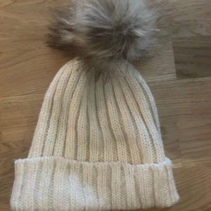 Ribbad vit stickad mössa med pälsboll - Snygg ribbad vit stickad mössa med stor fluffig pälsboll på toppen som inte är riktig päls. Perfekt för kalla dagar och ger en trendig look. Mössan har uppvikt kant och är riktigt mjuk och skön att bära.