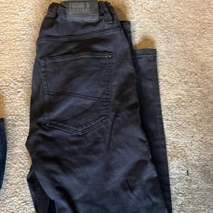 Svarta jeans från Young & Original - Svarta jeans med raka ben och slitna detaljer på låret och knät. Klassisk femficksmodell med en stor bakficka och diskret logga på linningen. Jeansen har en mörk ton och är tillverkade i ett mjukt bomullsmaterial som är skönt att bära.