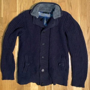 Pälsfodrad cardigan - Säljer en mörkblå stickad kofta från Ted Baker med grovstickad struktur och snygg krage fodrad med mjuk grå fleece. Koftan har knappar framtill, två fickor med knappdetaljer och långärmade ribbade muddar. Insidan har ett rutigt blått foder som ger extra stilpoäng. Är osäker vilken storlek det är men passar S/M