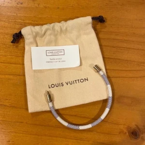 Louis Vuitton vitt armband med gulddetalj - Snyggt vitt armband från Louis Vuitton med ett cylinderformat guldfärgat lås där märket är ingraverat. Armbandet har ett diskret svart mönster längs hela bandet som ger en cool kontrast. Perfekt accessoar för dig som gillar lyxiga detaljer.